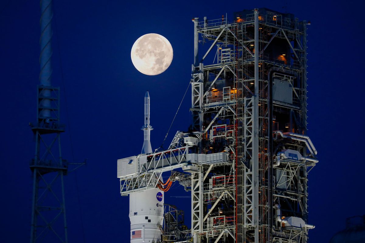 A rocket stands attached to a gantry. The full moon is right above the nose of the rocket.