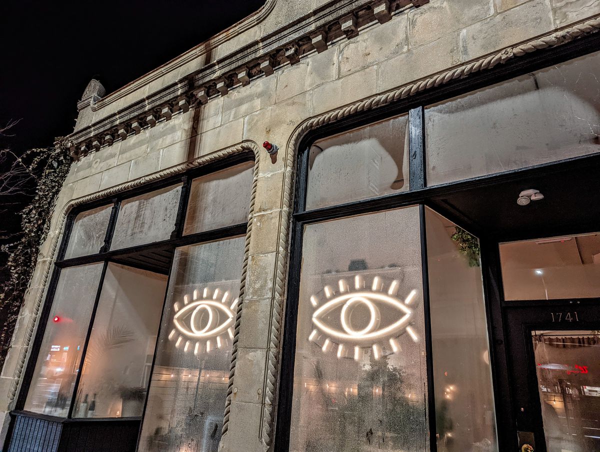 Two neon eyes light up the large front windows of a restaurant, which is built in a one-story stone building.