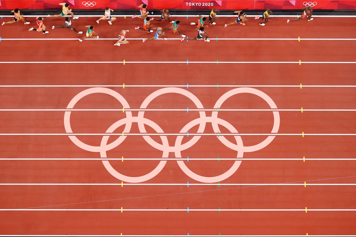The Olympic rings on a track and field track seen from high above, with competitors running in the inside lane.