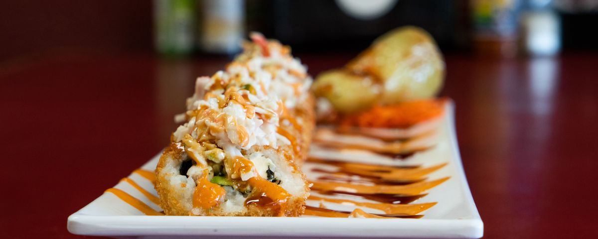 A fried sushi roll drizzled with orange sauce on a rectangular white plate. 