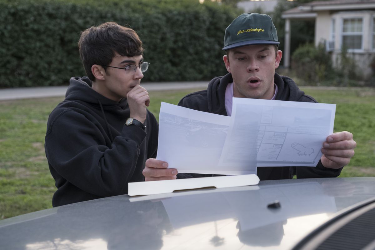 Jimmy Tatro, wearing a hoodie and a green baseball cap, looks stunned while reading documents on a car hood, while Tyler Alvarez looks on, biting his nails, in American Vandal.