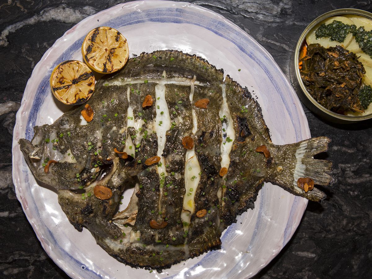 A large grilled whole fish on an intricate plate with a sides in a tin can.