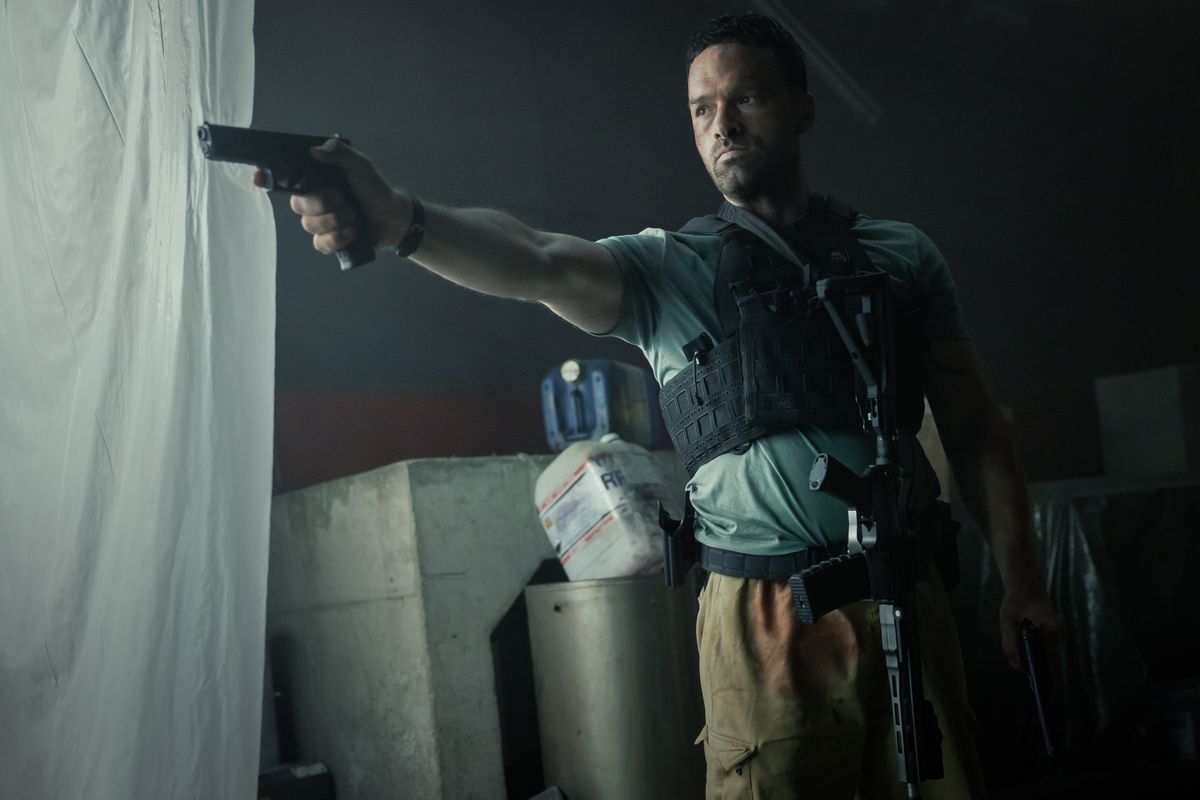 Alban Lenoir as Adam Franco, wearing a bullet-proof vest and a rifle hanging from his belt while holding a pistol in AKA.