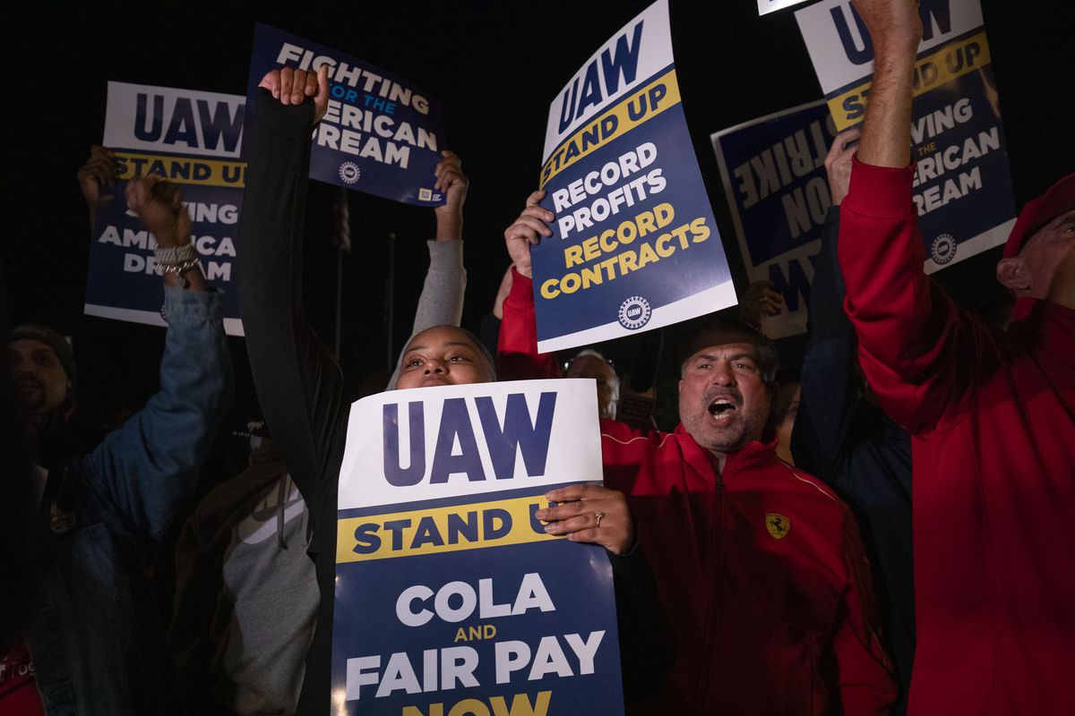 People on strike holding up signs saying “UAW stand up” and “fair pay.”