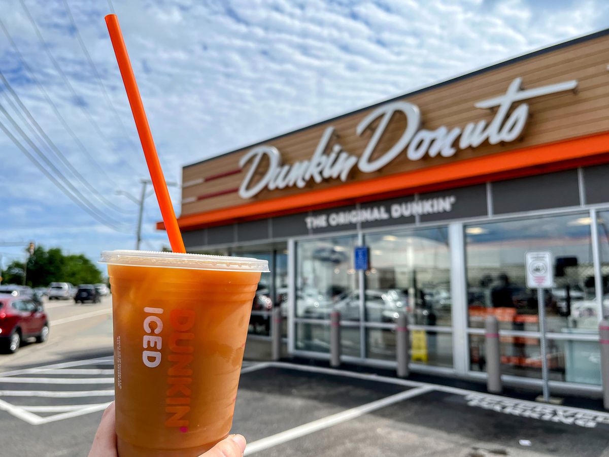 An iced coffee in the foreground with a retro-designed shop in the background. A sign is visible saying “Dunkin’ Donuts” and “The Original Dunkin’”