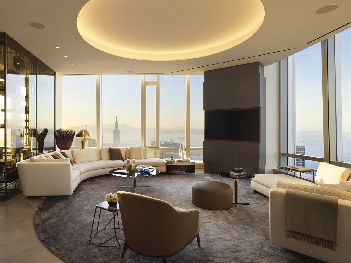 A living room with a circular indentation in the ceiling with recessed lighting inside, widows galore with views of the city and the fog, and half-circle couch and seating.