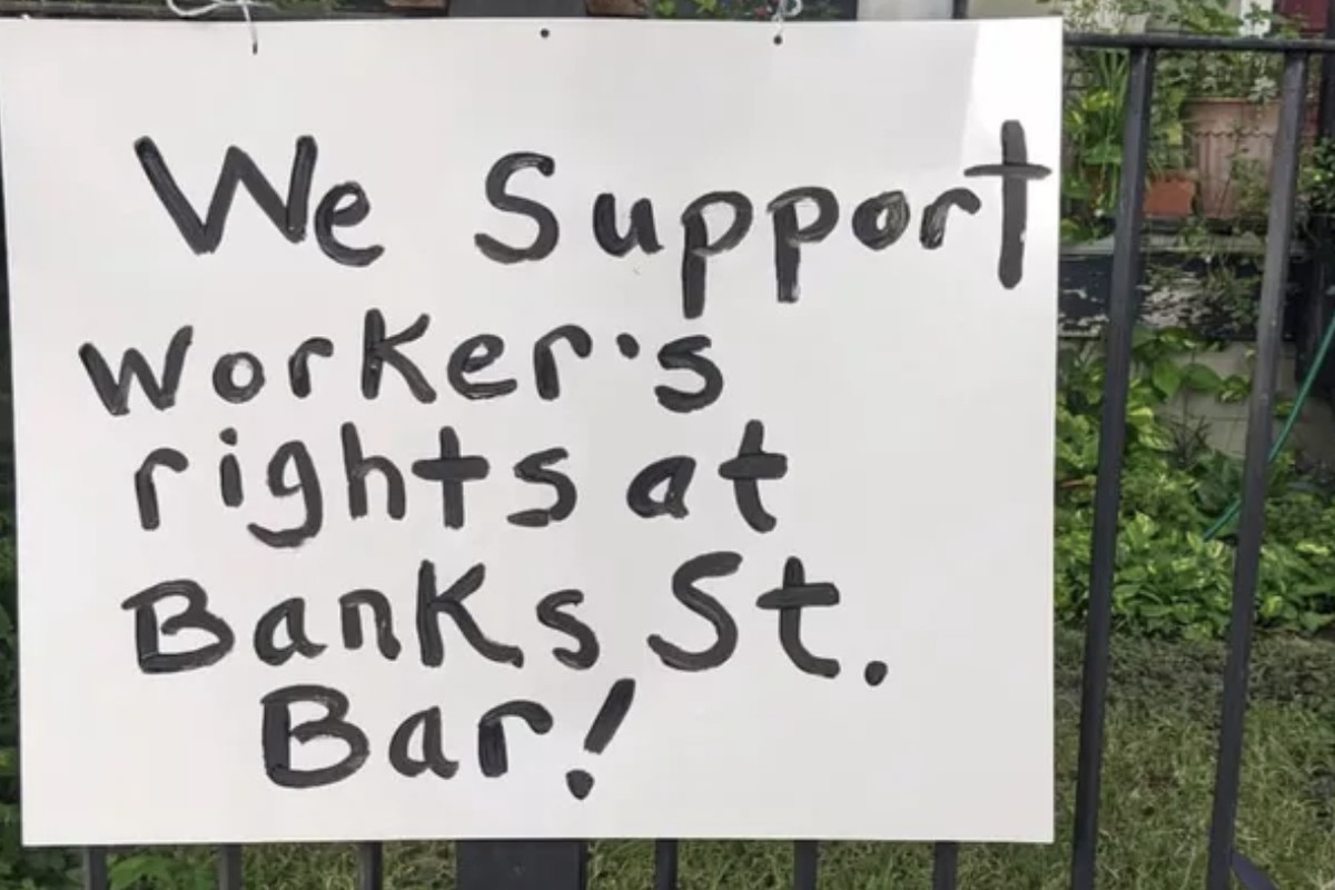 A sign reading “We support worker’s rights at Banks Street Bar” is attached to an iron fence.