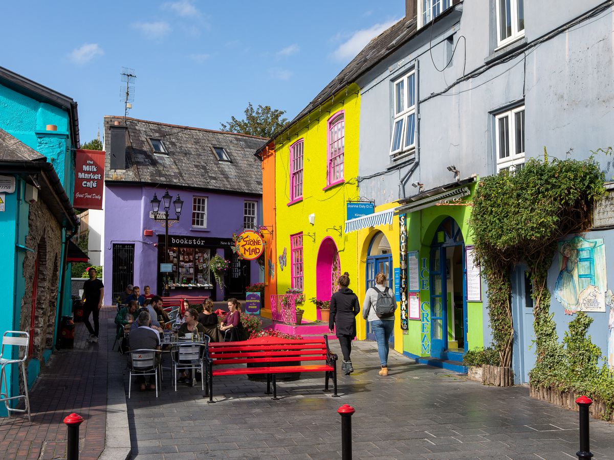 Colorful buildings and shops surround an outdoor patio on cobblestone streets, where customers sit in groups and others walk beneath a beautiful blue sky