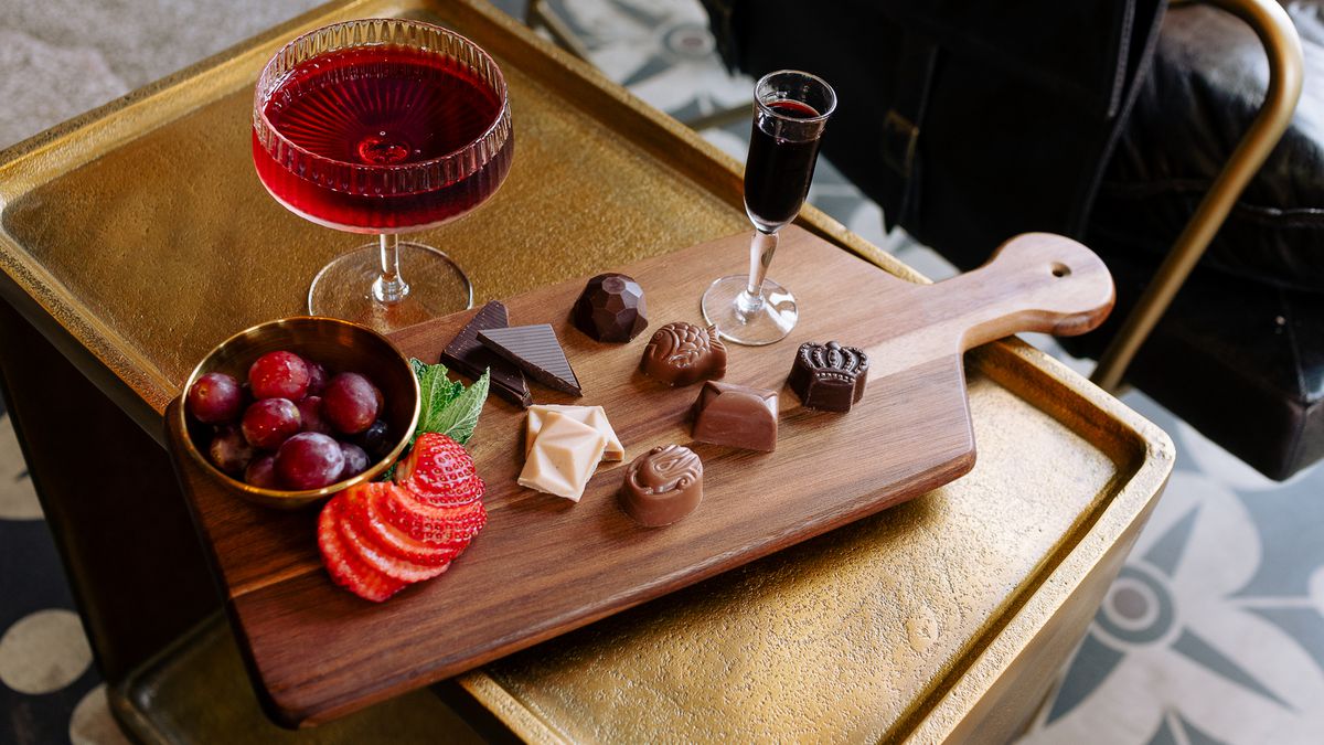 A gold tray holds a dark wood board, loaded with chocolates, fruits, a glass of port. Behind it, another port cocktail sits.