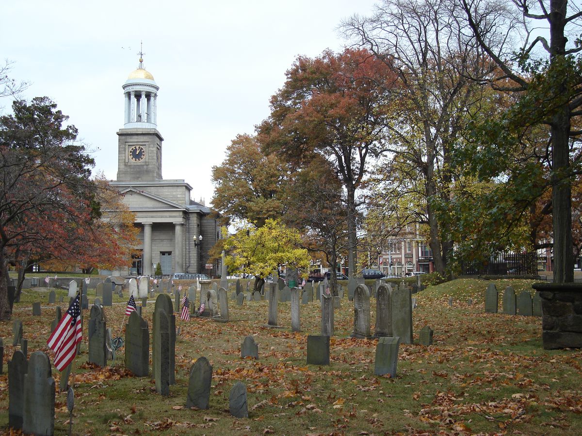 Rows of headstones amid leaf-covered ground, with a church with a spire in the distance.