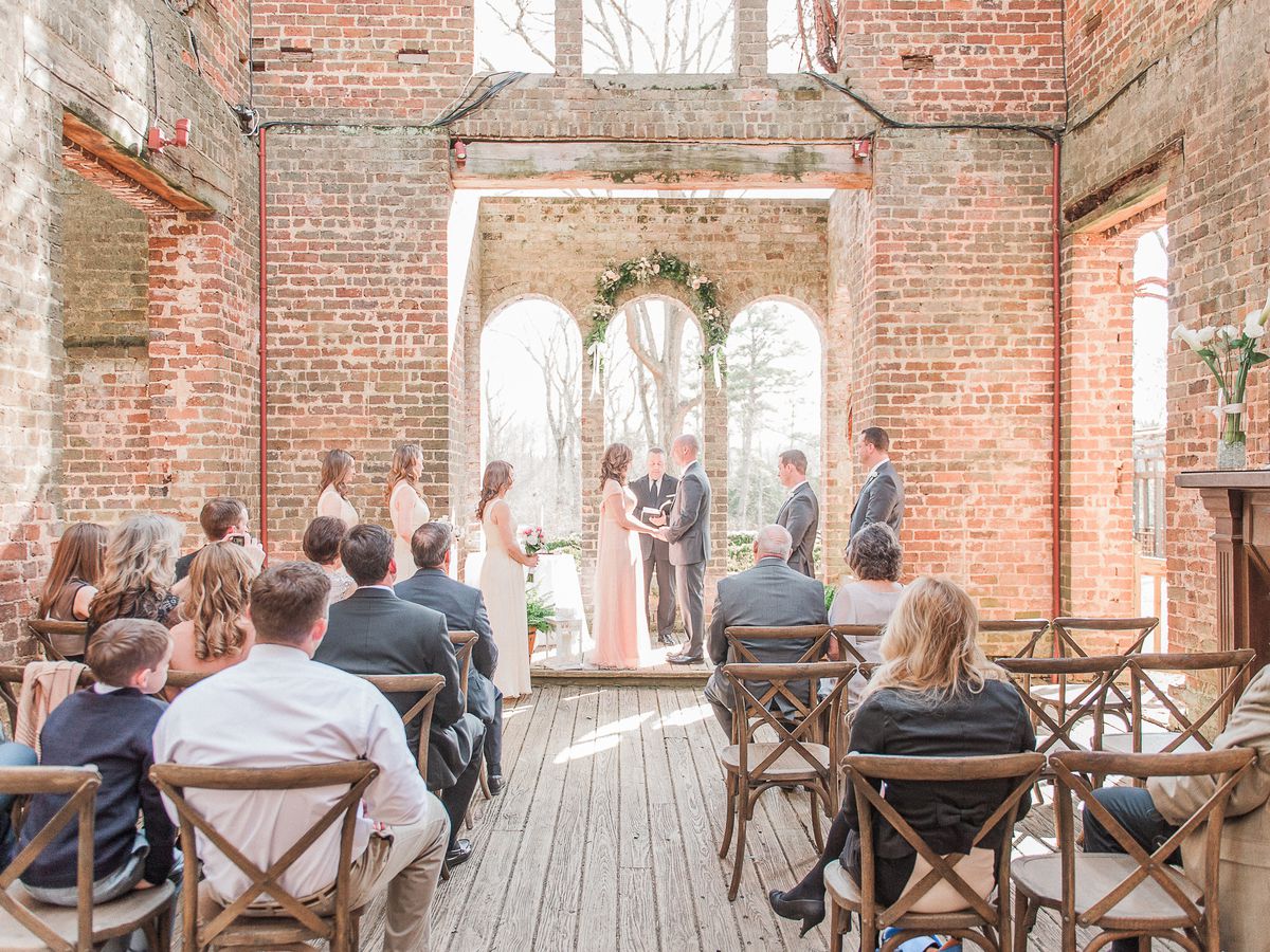 The interior of a wedding chapel. The walls are exposed red brick. There are chairs. There are people sitting on the chairs. There is a bride and groom standing at the altar in the front of the room.
