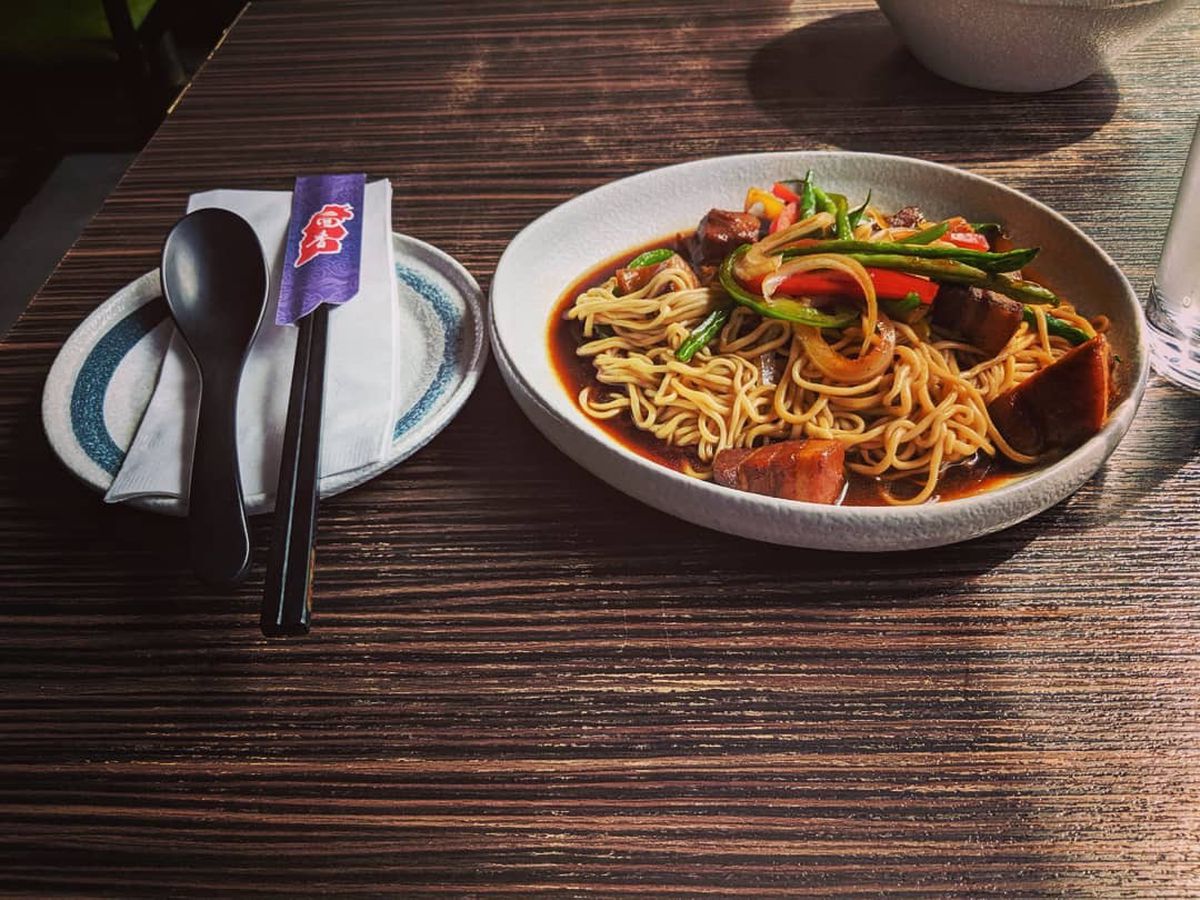 A table is set with a fork, spoon, and chopsticks on a saucer next to a large bowl of noodles topped with scallions, sliced bell peppers, in a dark broth
