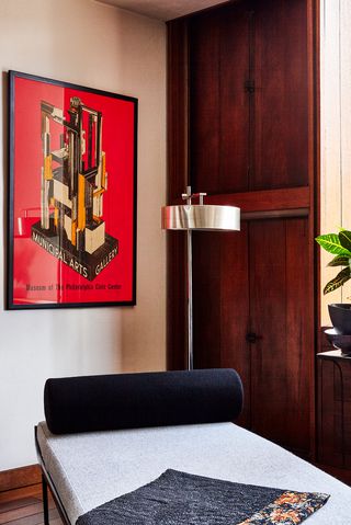 szara tapicerowana ławka z czarną poduszką na szyję, minimalistyczna Srebrna Lampa Podłogowa i plakat retro z abstrakcyjną ilustracją na czerwonym tle