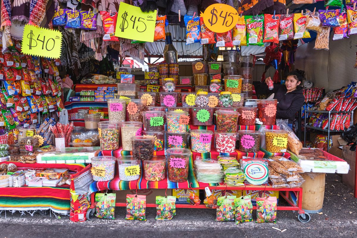 A large market stall displays deep shelves stacked high with various chile-covered candies and gummy snacks. A diverse and broad array of bagged chips, peanuts, and more cover the stall’s side wall and hang from a clothesline above the shelves.