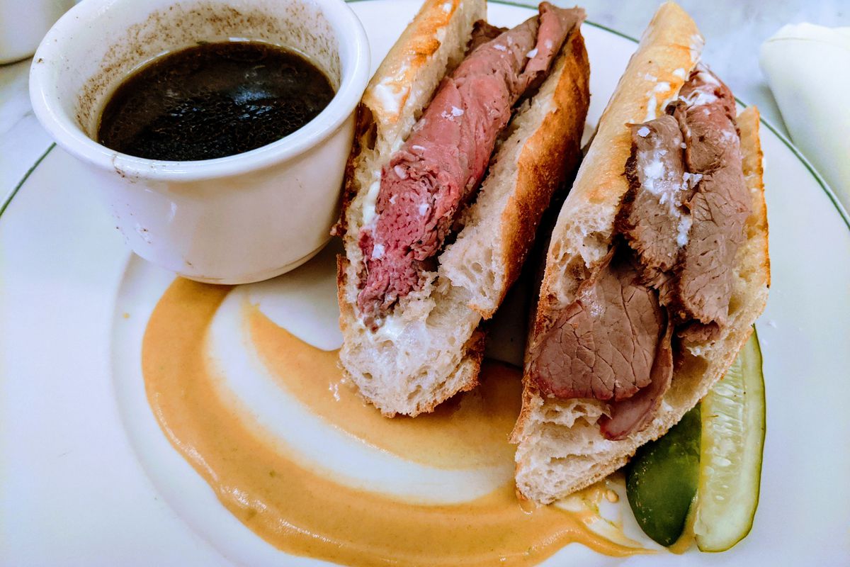 Two halves of a roast beef hero on a baguette with dark broth on the side.