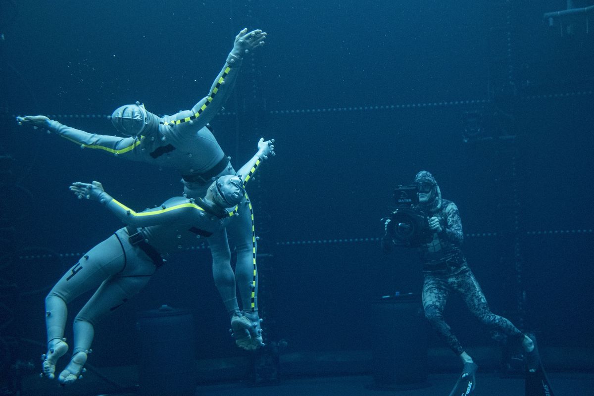 An underwater motion capture technician holds a camera-like device aimed at two actors swimming around with their arms outstretched in a pool for Avatar: The Way of Water