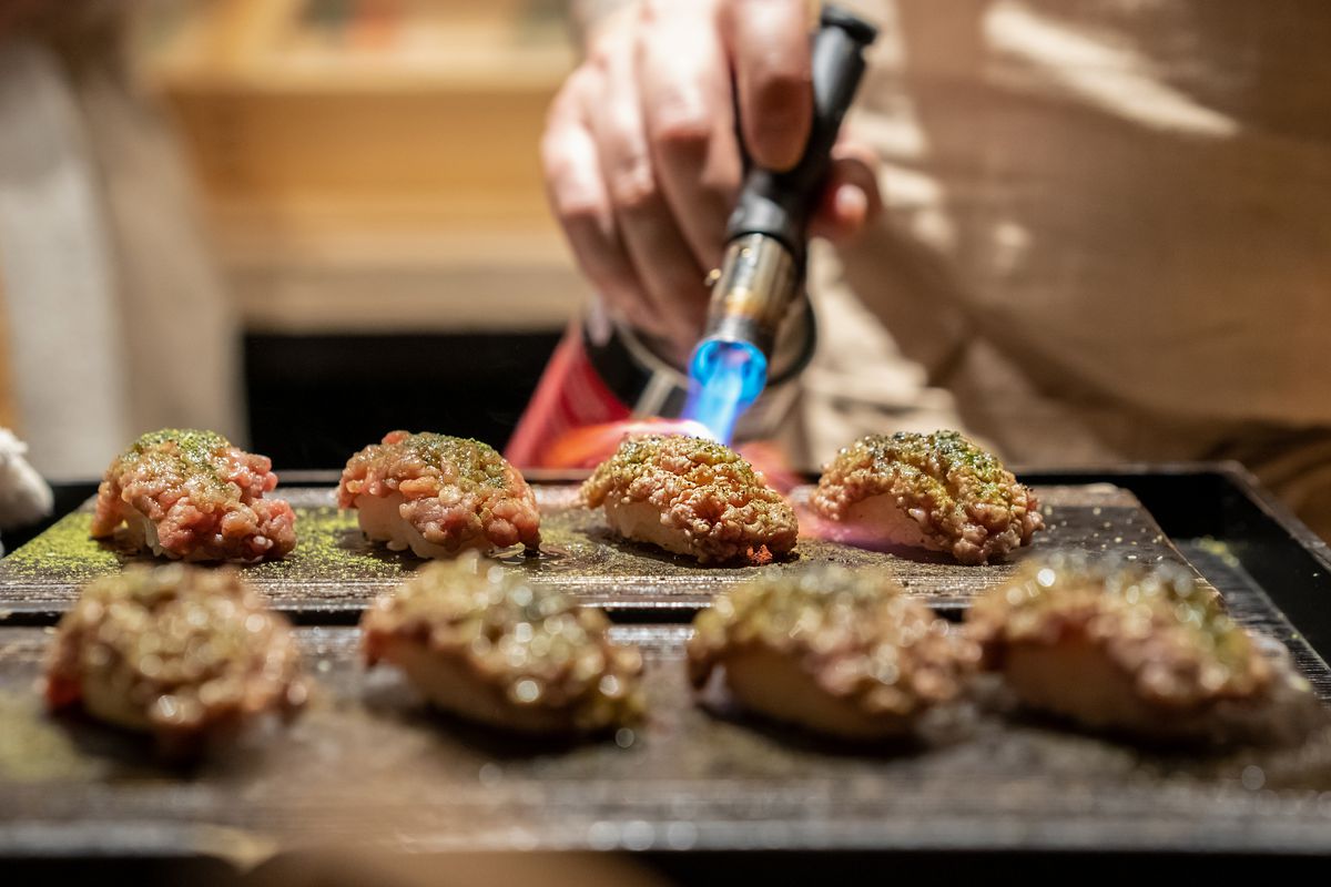 A person blow-torching a piece of meat-covered sushi on a tray of meat-covered sushi pieces.