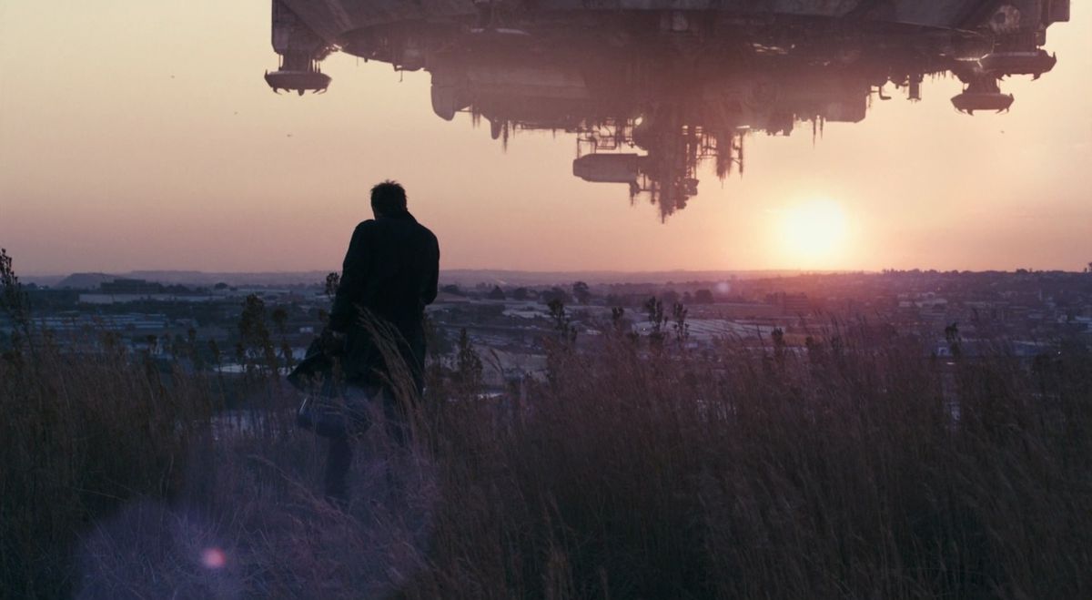 Sharlto Copley in a raincoat standing in a field overlooking a large spaceship flying in the sky as the sun sets in District 9.