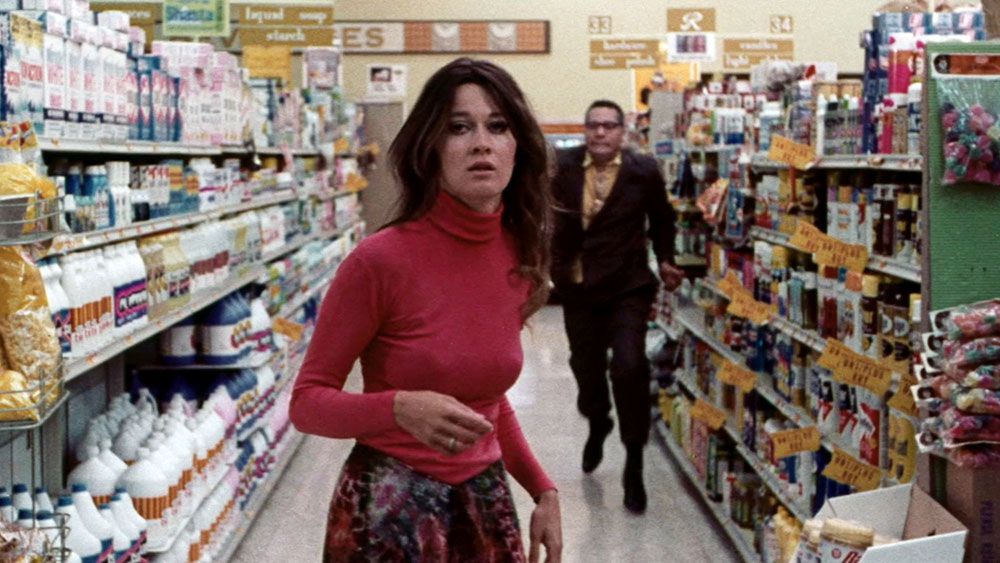 A distressed woman in a red sweater stands unsuspectingly in a grocery store aisle as a man in a brown suit stalks behind her in Messiah of Evil.