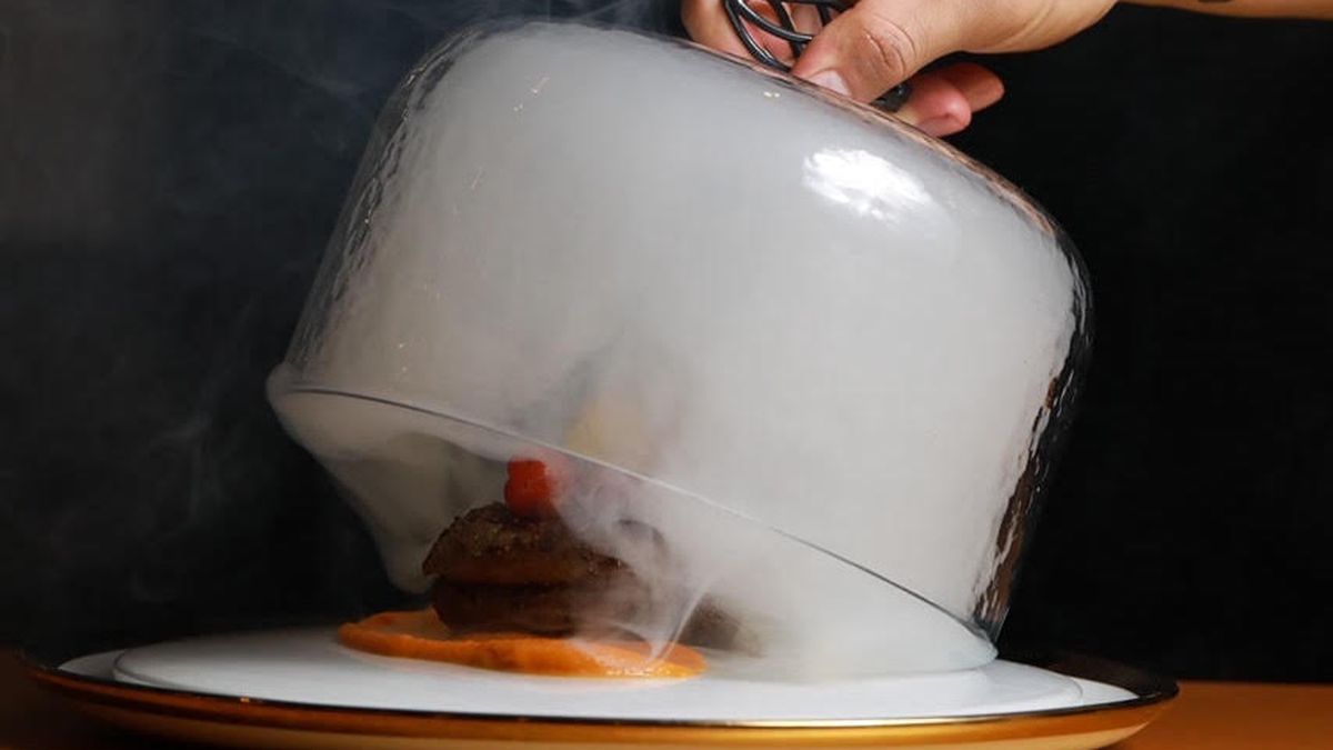 A hand reaches in to uncover a glass topper, revealing smoke and a steak with tomatoes on top, at new LA restaurant Juliana.
