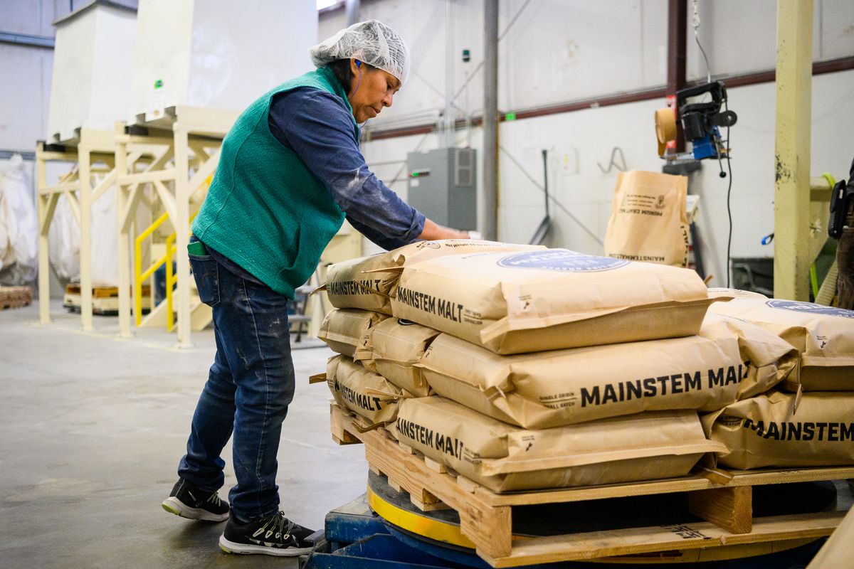 A woman in a hair net fills bags of malt next to filled bags on a pallet.