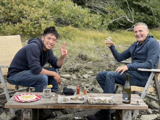 A Japanese American man sits in a chair, flashing a peace sign, across from an older man who is raising his wine glass. There is a spread of food in front of them.