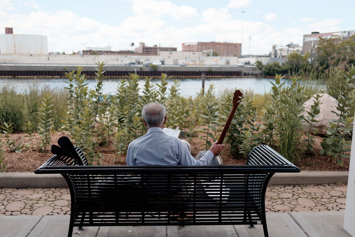 A man holding a Chinese-stringed musical instrument while enjoying the day sitting on a riverside bench.