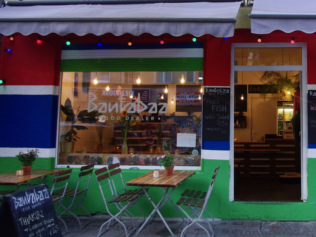 A colorful restaurant exterior, with large bans of red, green, and blue paint beneath an awning, with a few outdoor tables and chairs