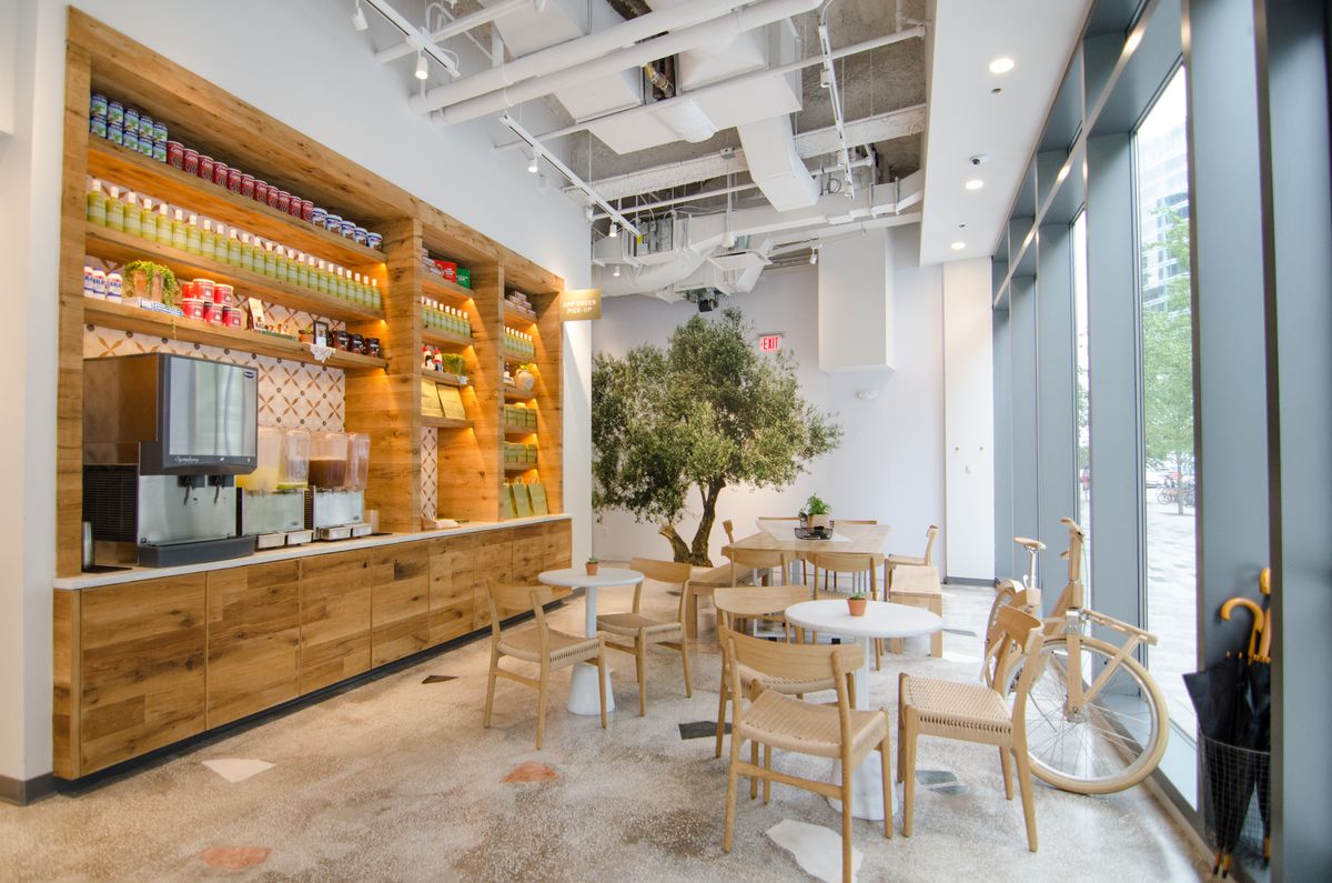 One portion of Greco Seaport includes tables, an olive tree mural, umbrellas, a wooden bicycle, beverage dispensers, and a variety of Greek products on shelves