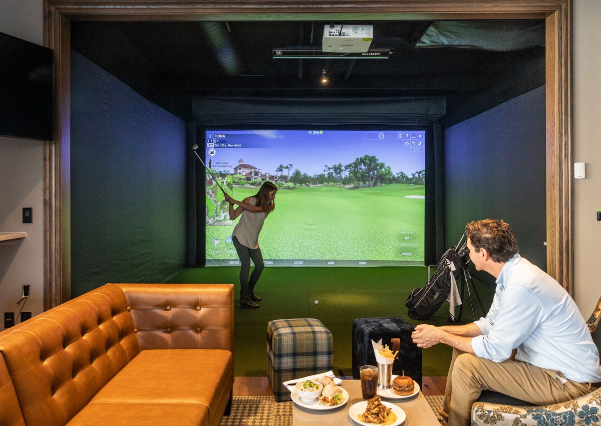 A women swings into a screen with a golf course simulated on it. A man watches her from a couch