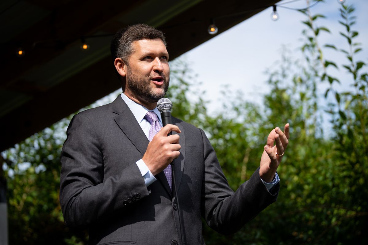 Ryan, dark-haired in a gray suit and purple tie, speaks on an outdoor stage, backed by green trees on a sunny day.