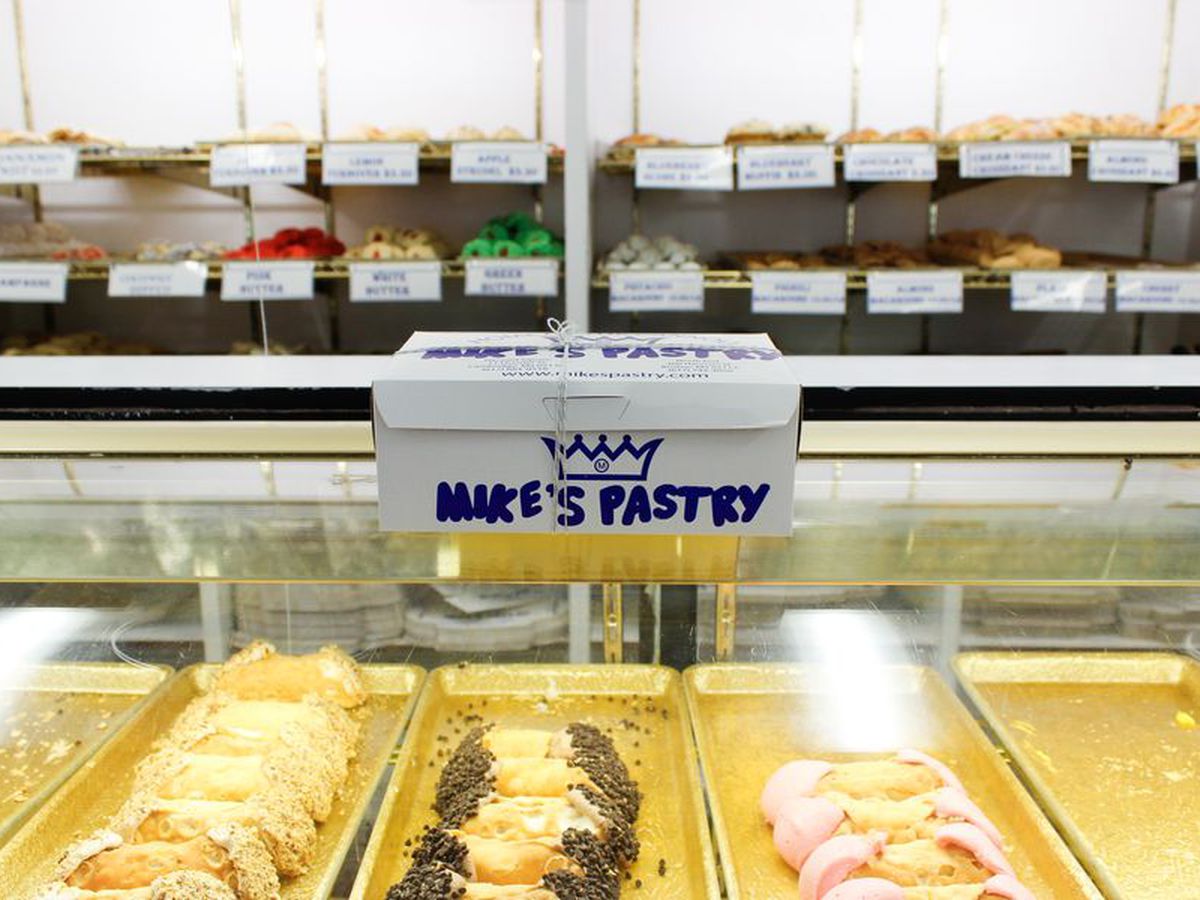 A pastry case in an old Italian shop features several rolls of cannolis. A white box with blue text that reads Mike’s Pastry sits atop the case.
