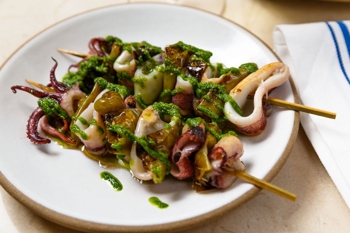 Two skewers stacked with squid and green peppers and drizzled in green oil lay side by side on a white plate