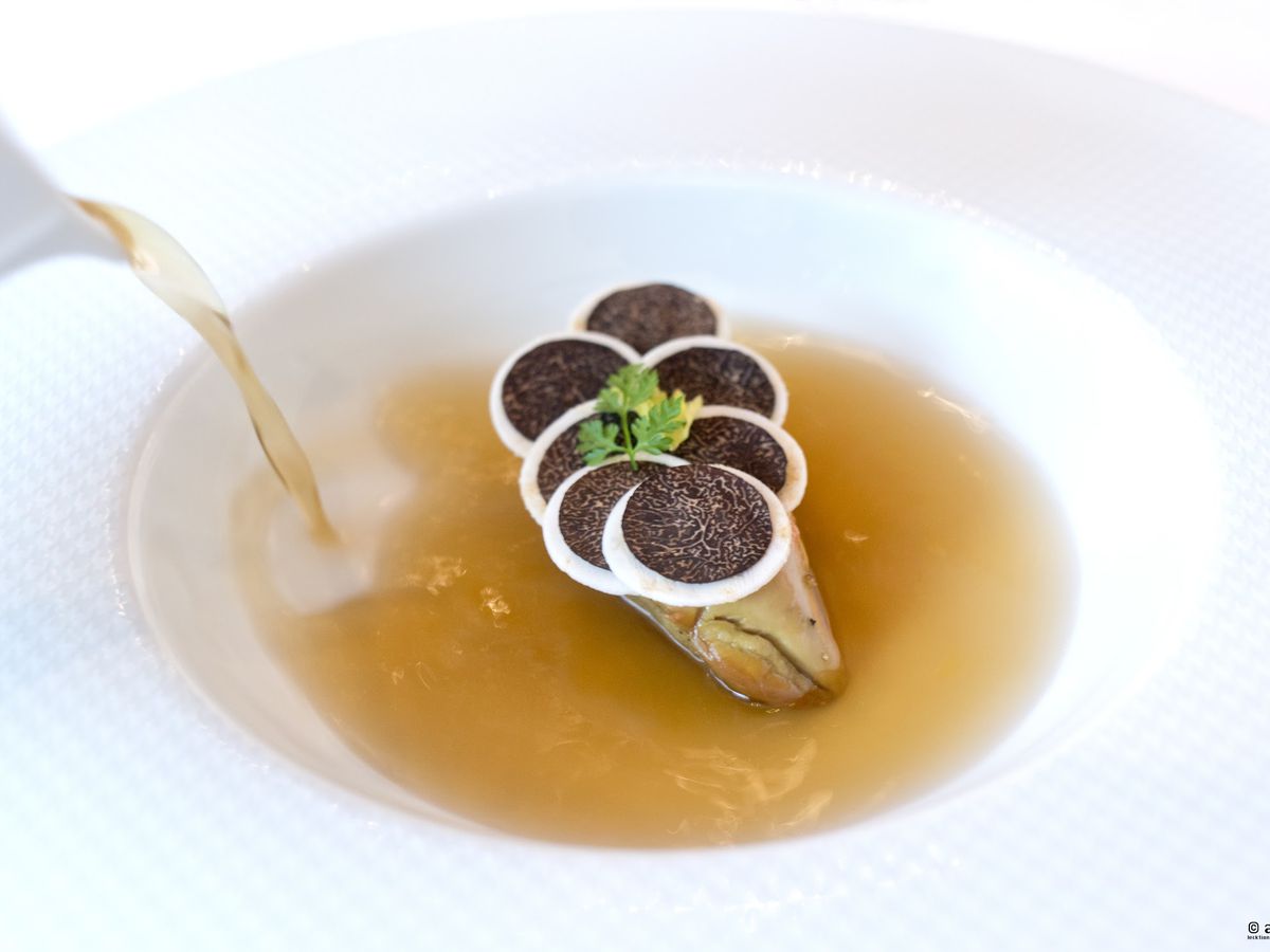 An unseen server pours consomme into a bowl around a mound of foie gras.