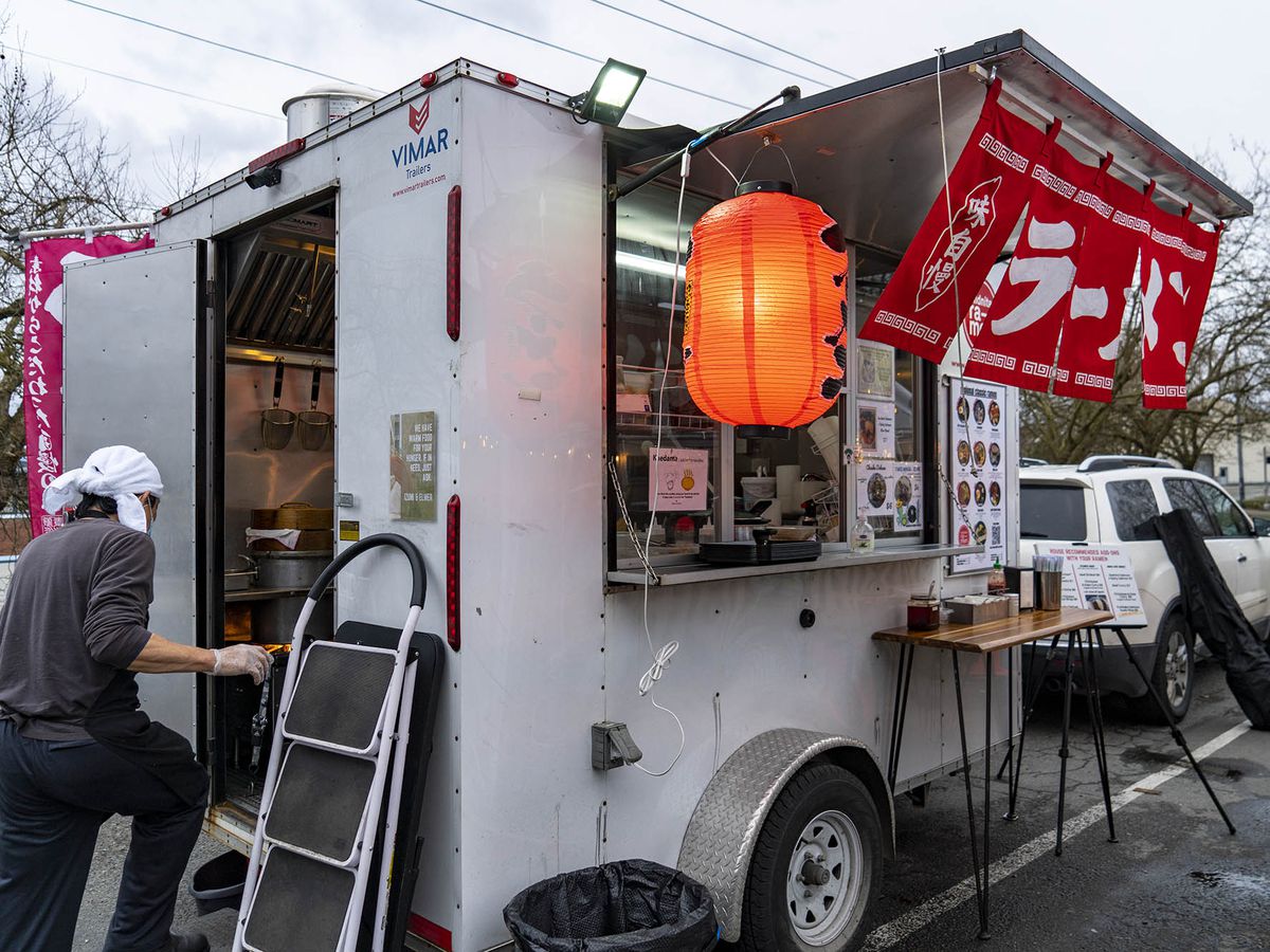 A man wearing a white head scarf enters a small mobile food cart with red flags displaying Japanese characters and an orange glowing paper lantern out front.
