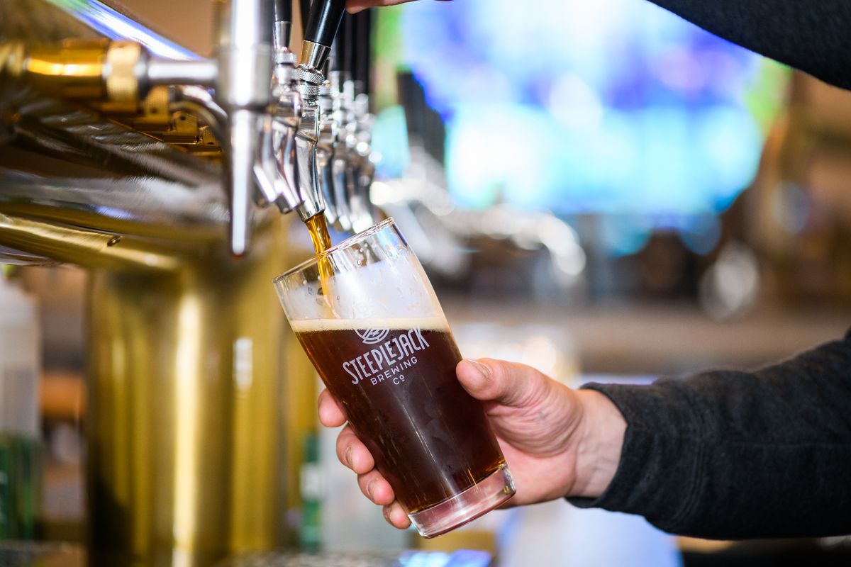 A white hand holds a pint glass filled with a dark, coffee-brown beer, as he pours it from the brewery’s taps.