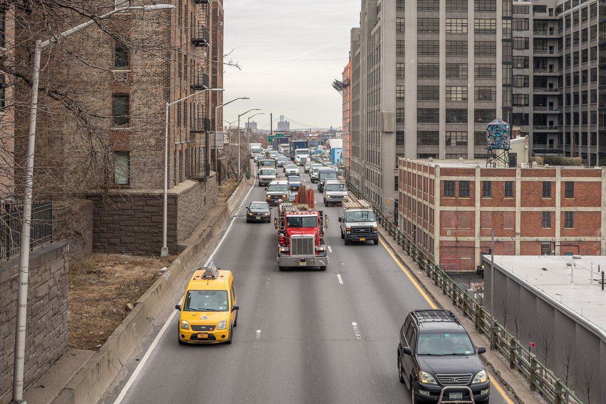 A yellow cab, a red truck, and several other cars driving on the Brooklyn Heights section of the Brooklyn Queens Expressway.
