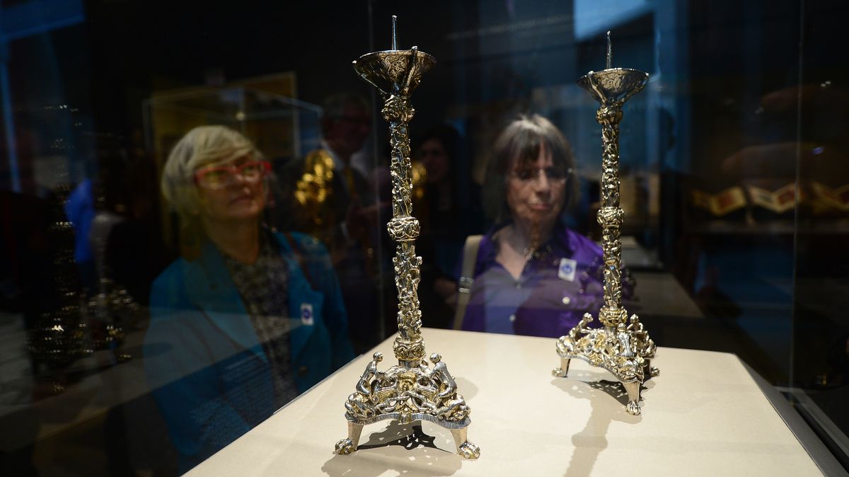 Two ornate candlesticks in a glass display case, observed by two museum-goers.