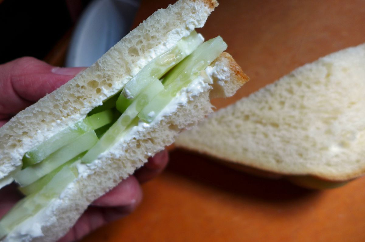 A hand holds a half sandwich cut to show cucumber and white cream cheese on white bread.