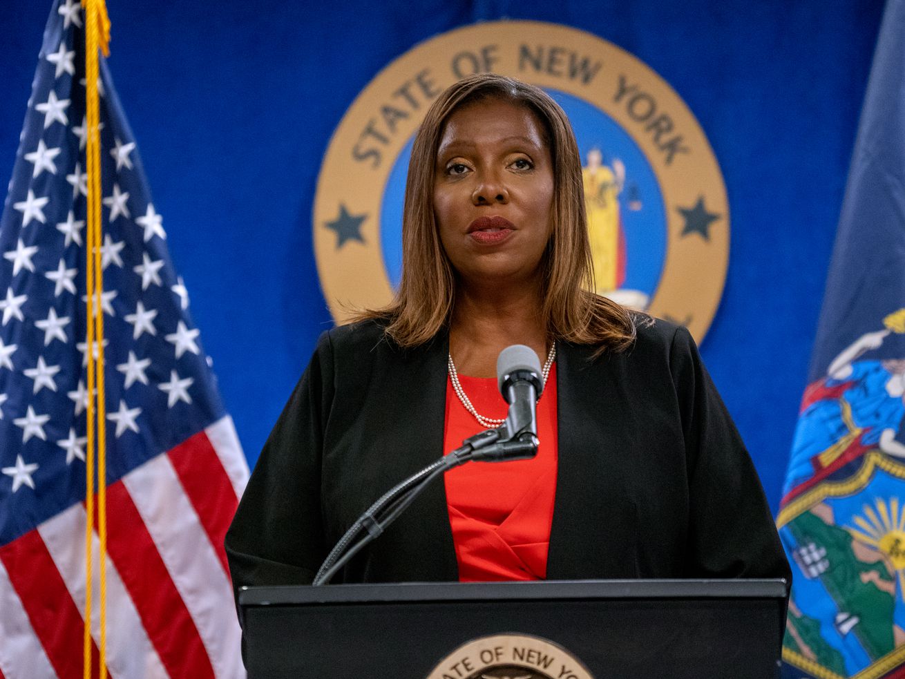New York Attorney General Letitia James standing at a lectern and speaking into a microphone, with the seal of the state of New York behind her and flags on either side.