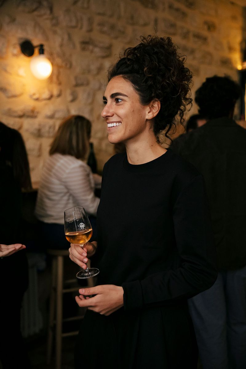 A woman in all black smiles while holding a wine glass.