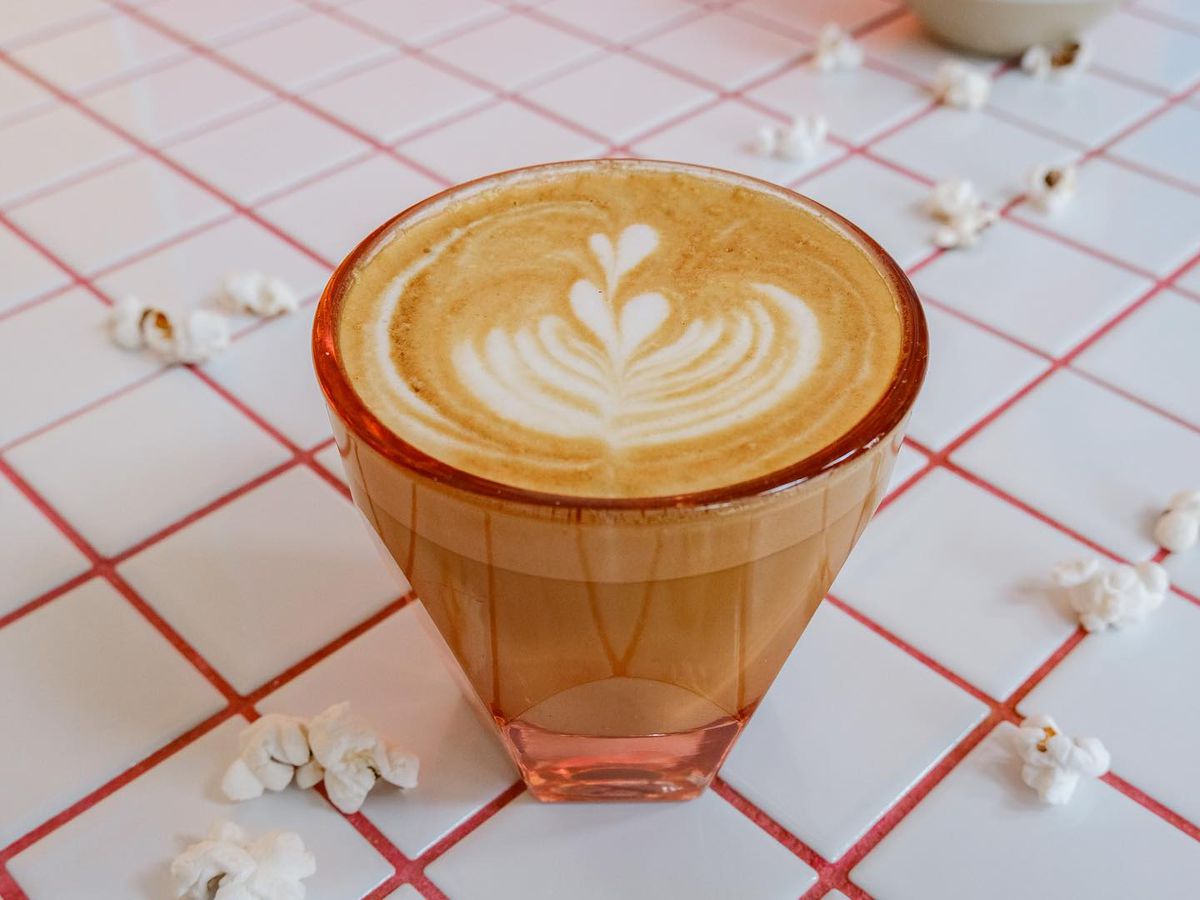 A cup of a coffee in a clear red glass with latte art surrounded by popcorn kernels