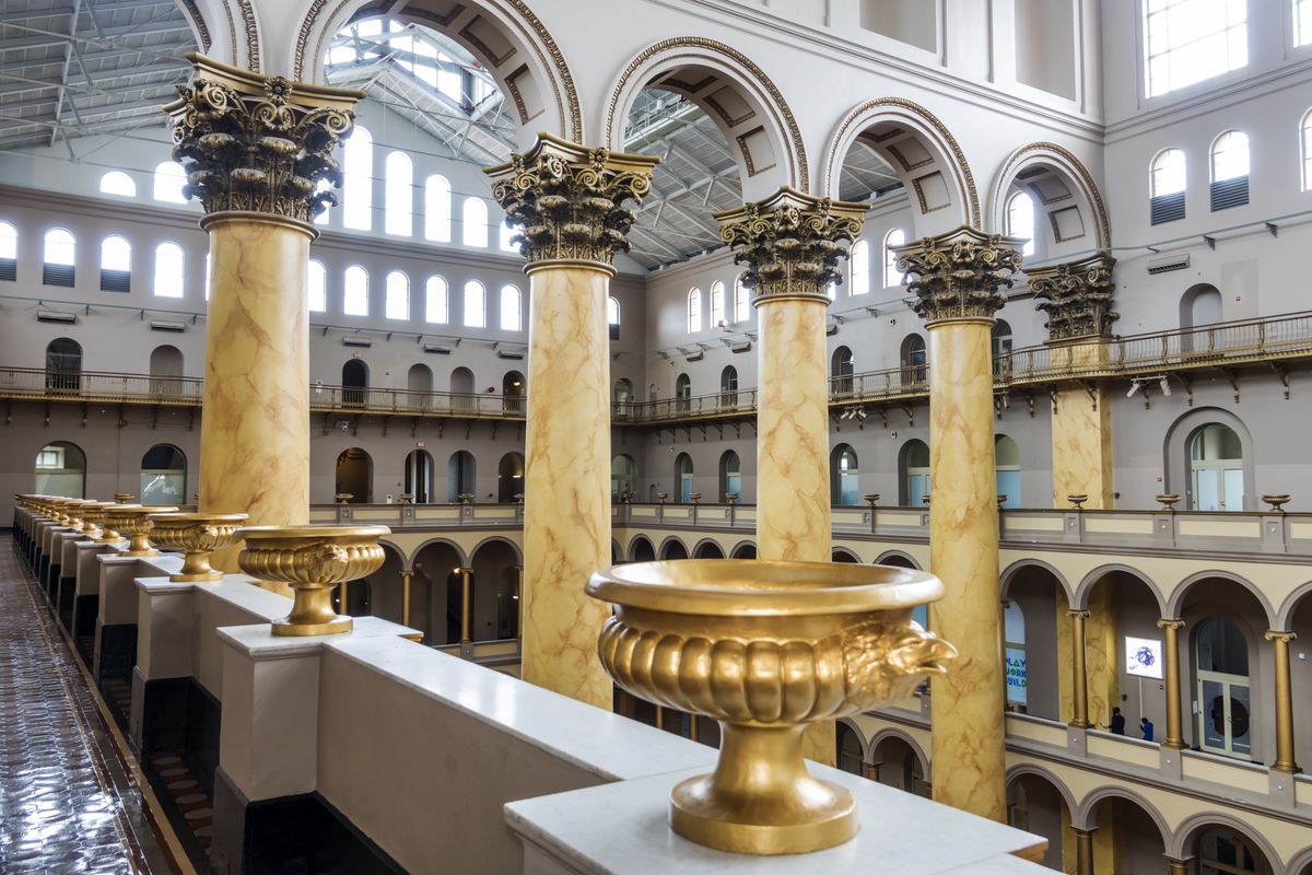 A row of Corinthian columns inside a grand romanesque building. There are gold urns along a balcony.