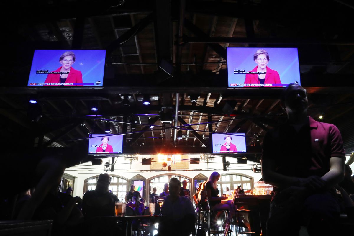 Senator Elizabeth Warren appears on several video screens above a crowd gathered at a bar.