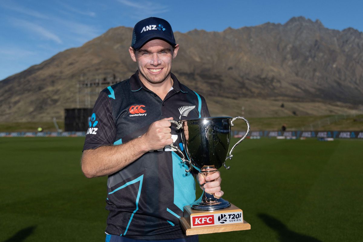A cricket player, in a grey jersey with aquamarine highlights. holding a large cup style trophy with curved highlights
