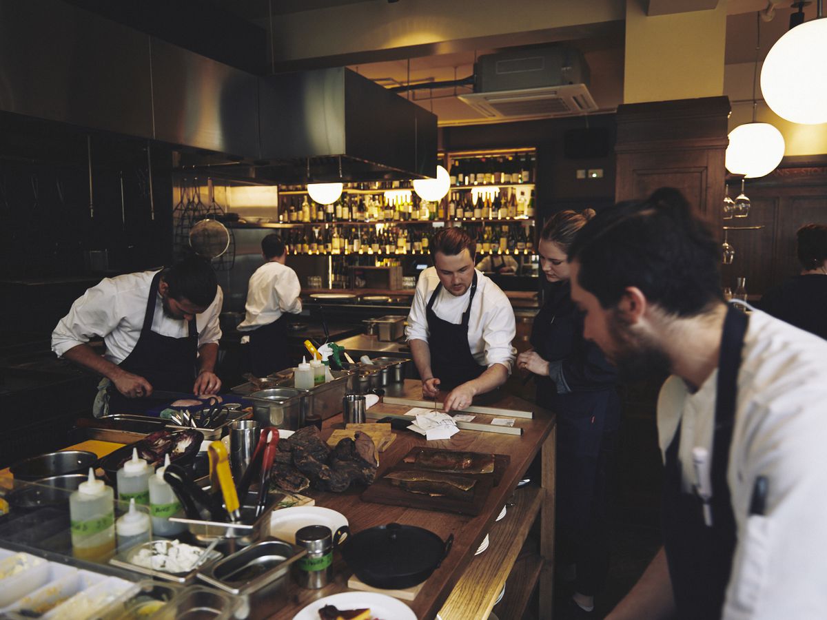 Chefs stand and work in blue aprons and whites at the pass at Brat restaurant Shoreditch