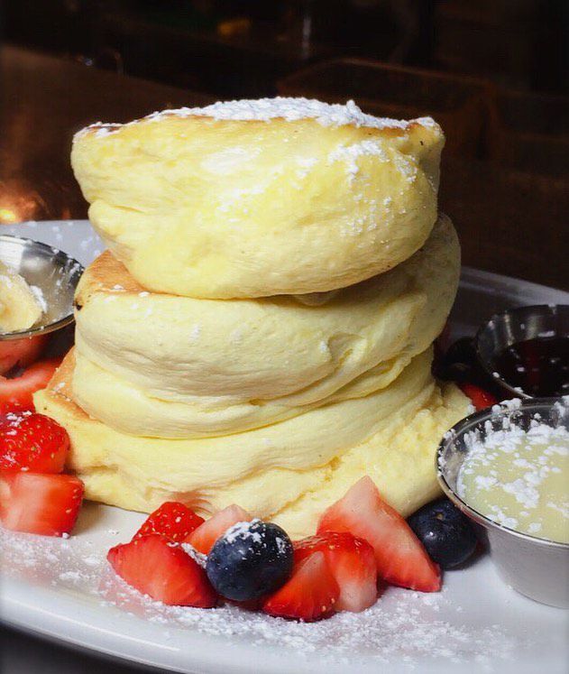 A stack of very thick souffle-style pancakes sit on a plate with a garnish of blueberries, strawberries, and powdered sugar