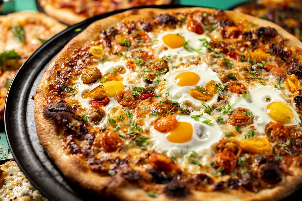 A pizza holds fried eggs, bacon, and roasted cherry tomatoes.