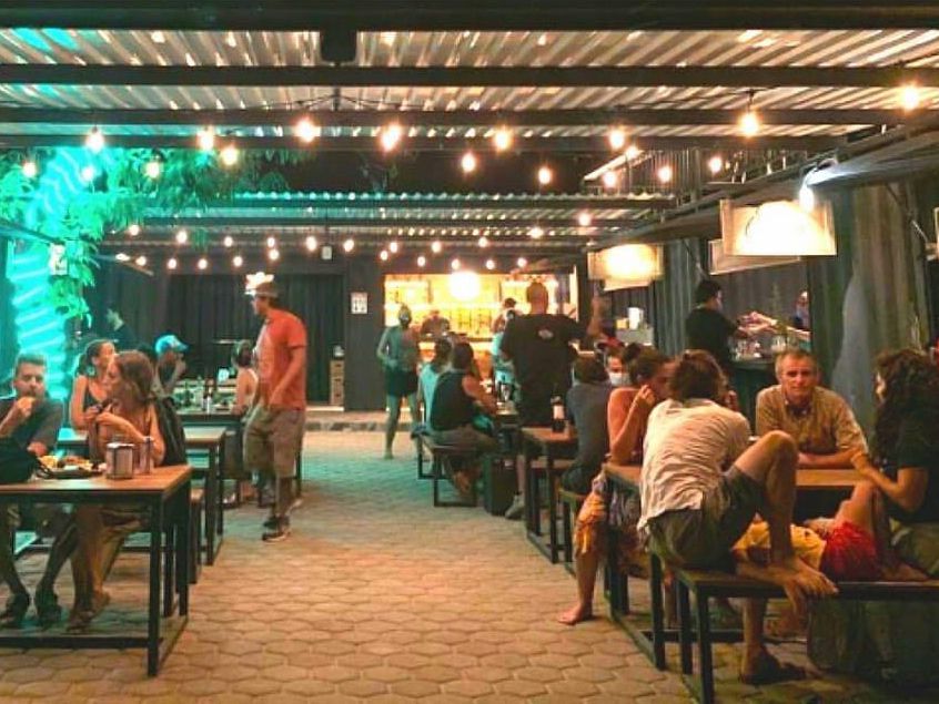 Diners sit at picnic tables in a metal-roofed space lit by string lights and bright blue neon glowing on indoor plants, with a kitchen visible in the background