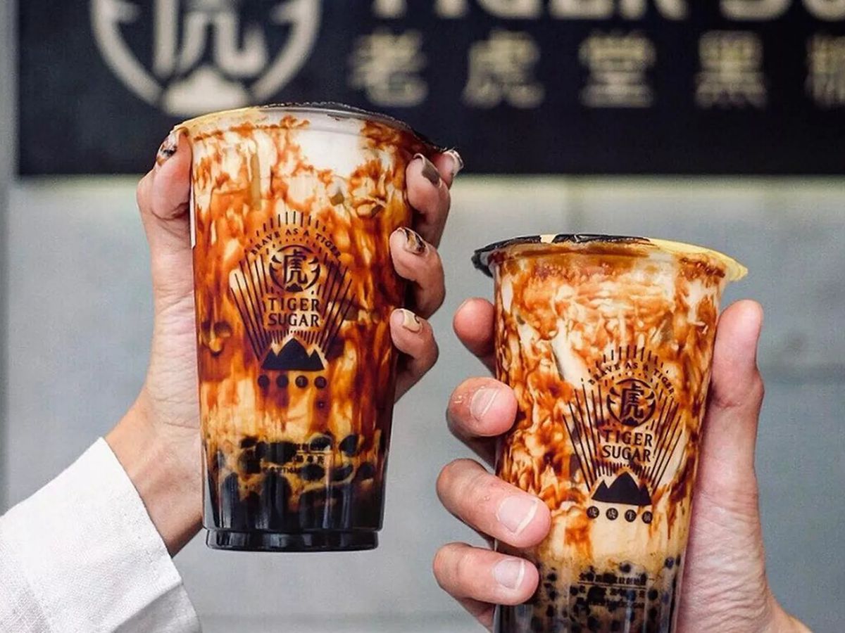 Two hands hold cups of brown sugar boba from Taiwanese chain Tiger Sugar. The iced tea drink is milky white with streaks of dark brown.